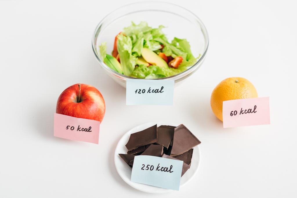 Friss gyümölcsök, csokoládé és saláta kalóriával kártyán fehér alapon, kalóriaszámláló diéta - Fotó, kép