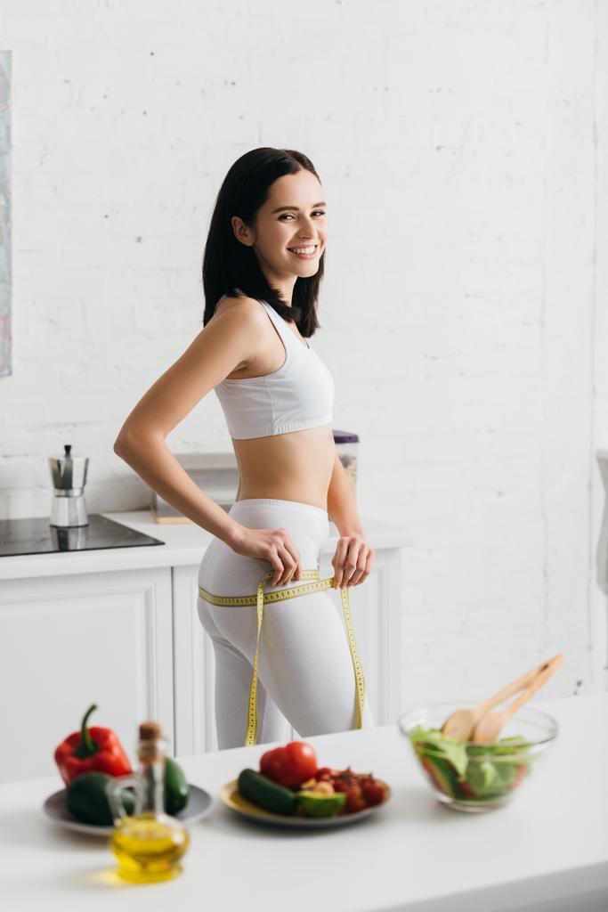 Selective focus of beautiful sportswoman smiling at camera while measuring hips with tape near salad and vegetables on kitchen table  - Photo, Image