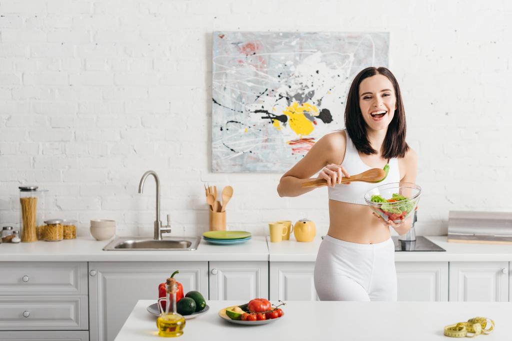 Beautiful sportswoman smiling at camera while holding bowl with salad near ripe vegetables and measuring tape on kitchen table - Photo, Image
