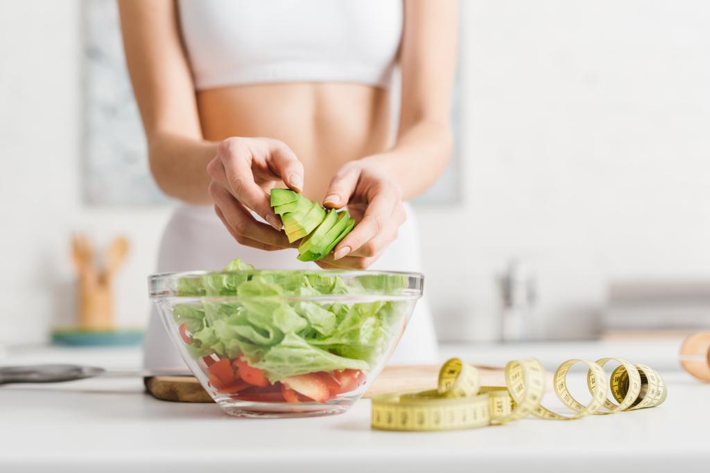 abgeschnittene Ansicht einer schlanken Frau, die Salat mit frischem Gemüse und Avocado in der Nähe von Maßband auf dem Küchentisch kocht - Foto, Bild