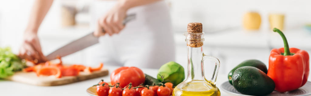 Selektivní zaměření olivového oleje a čerstvé zeleniny v blízkosti ženy vaření salát na kuchyňském stole, panoramatický výstřel - Fotografie, Obrázek