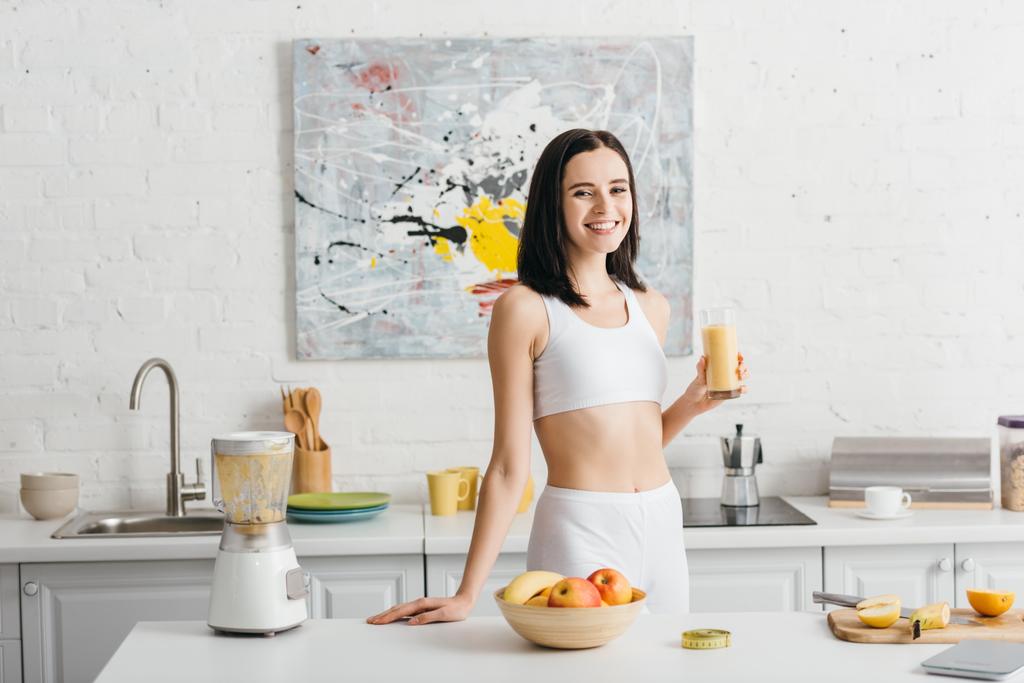 Çekici sporcu kadın mutfakta bir bardak smoothie içerken kameraya gülümsüyor, kalori sayıyor. - Fotoğraf, Görsel