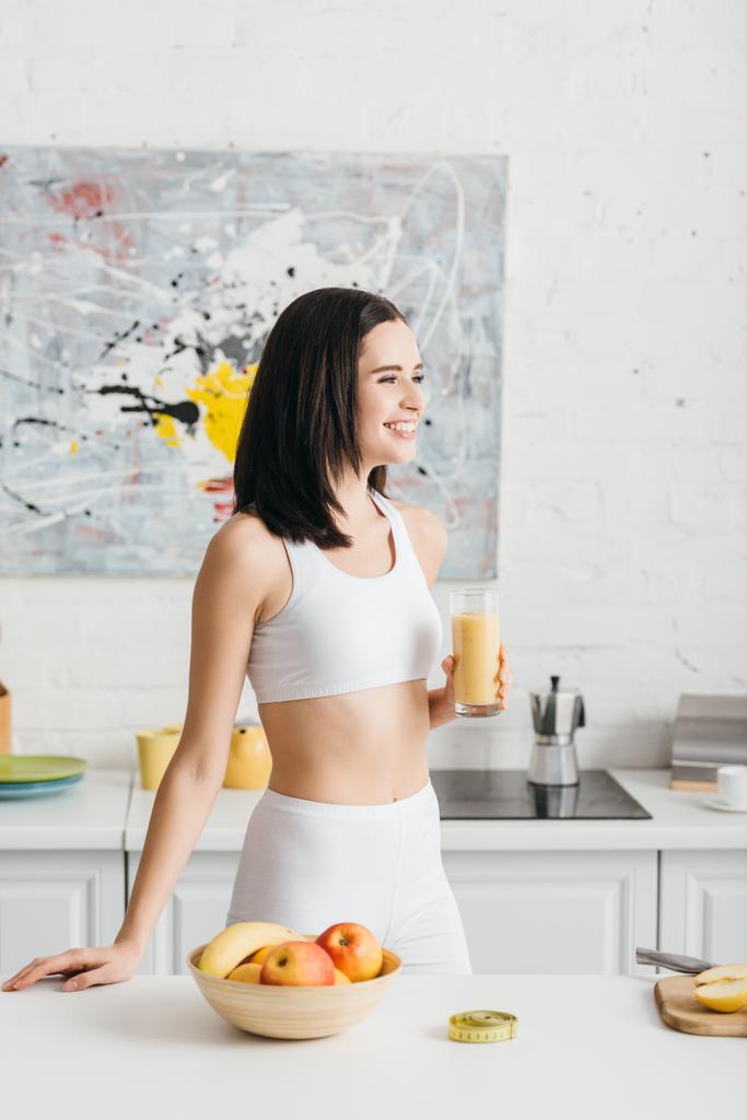 Meyvelerin yanında gülümseyen ve mutfak masasındaki şeritleri ölçen bir bardak smoothie ile zayıf bir sporcu kadın. - Fotoğraf, Görsel