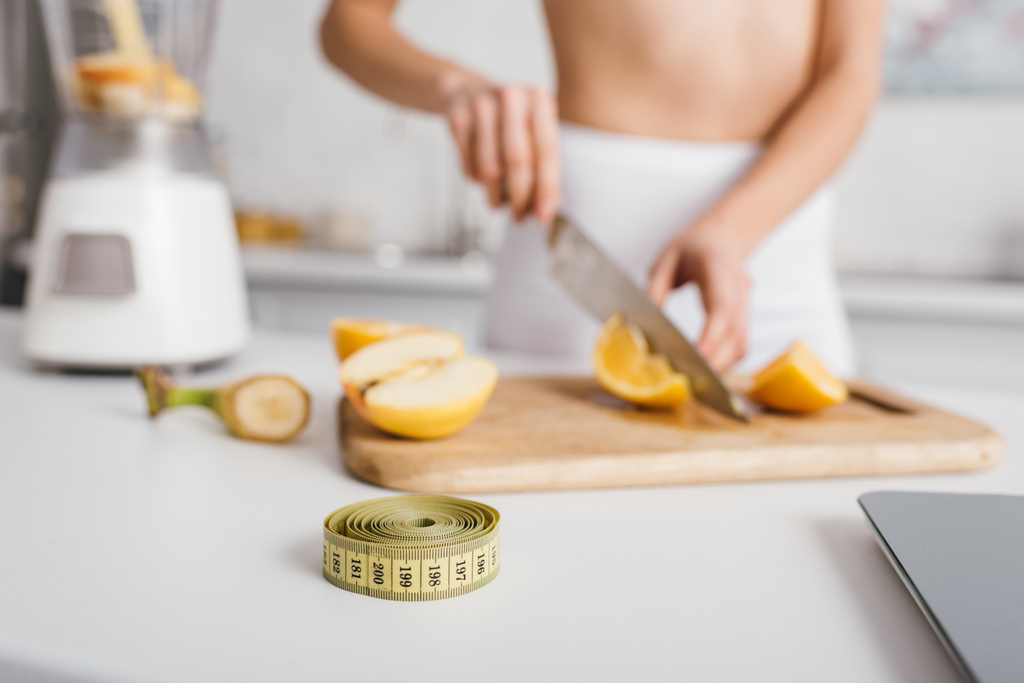 Selective focus of measuring tape and scales near fit girl cutting fresh vegetables for smoothie on kitchen table, calorie counting diet - 写真・画像