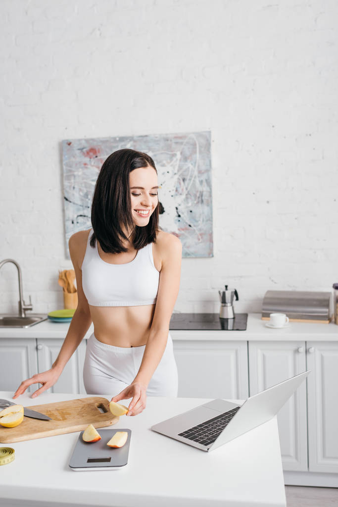 Beautiful smiling sportswoman looking at laptop while cutting apple near scales and measuring tape on kitchen table, calorie counting diet - Photo, Image