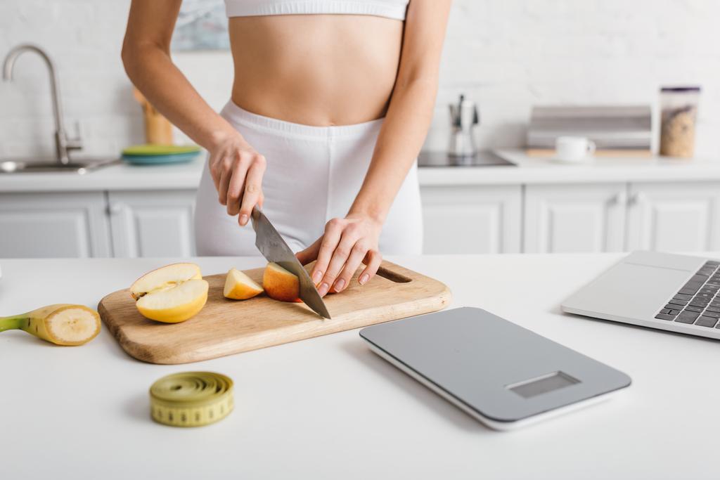 Cropped view of sportswoman cutting fruits near laptop, scales and measuring tape on kitchen table, calorie counting diet - Fotoğraf, Görsel