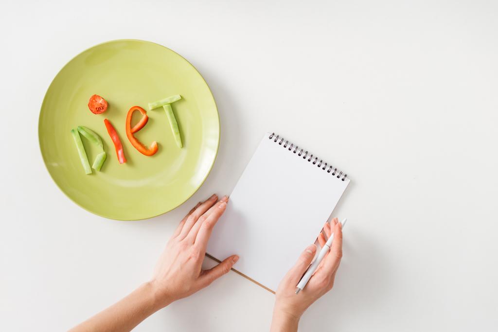 Top view of woman with pen and notebook near plate with diet lettering from vegetable slices on white background - Photo, Image