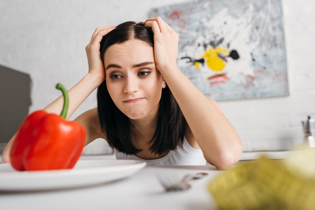 Selective focus of pensive woman looking at bell pepper near measuring tape on table - 写真・画像