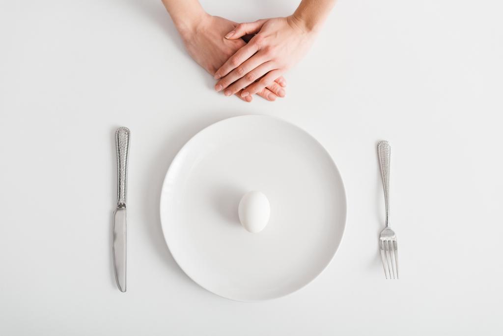 Vista dall'alto delle mani femminili vicino all'uovo su piatto e posate su sfondo bianco
 - Foto, immagini