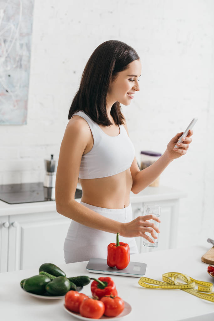 Zijaanzicht van lachende sportvrouw met smartphone en glas water in de buurt van groenten en meetlint op tafel, calorieën tellen dieet - Foto, afbeelding