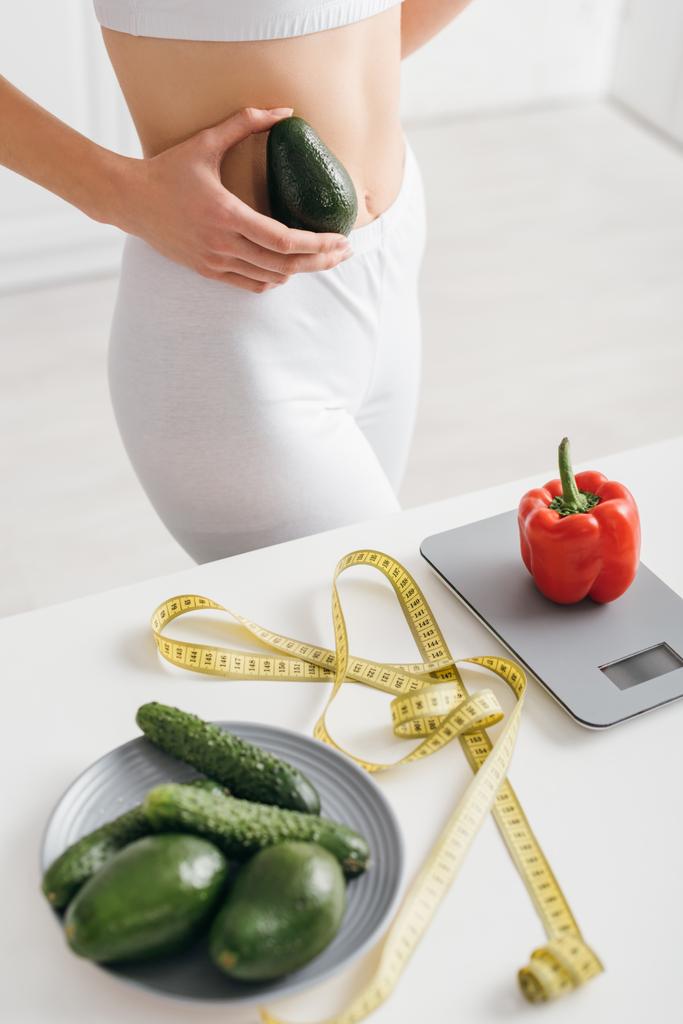 Обрезанный вид пригодной спортсменки, держащей авокадо рядом с перцем на весах и измеряющей лентой на кухонном столе, диета для подсчета калорий
 - Фото, изображение