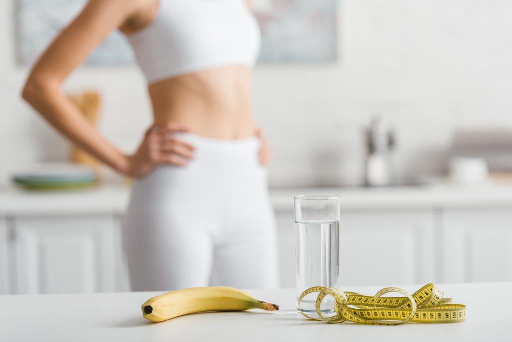 Селективный фокус банана, стакан воды и измерительная лента на столе рядом со спортсменкой на кухне
  - Фото, изображение