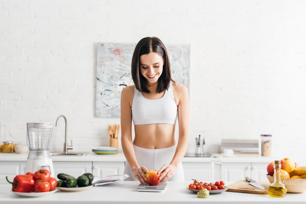 Belle sportive souriante pesant la pomme près des légumes, ruban à mesurer et carnet sur la table de cuisine, régime de comptage des calories
 - Photo, image
