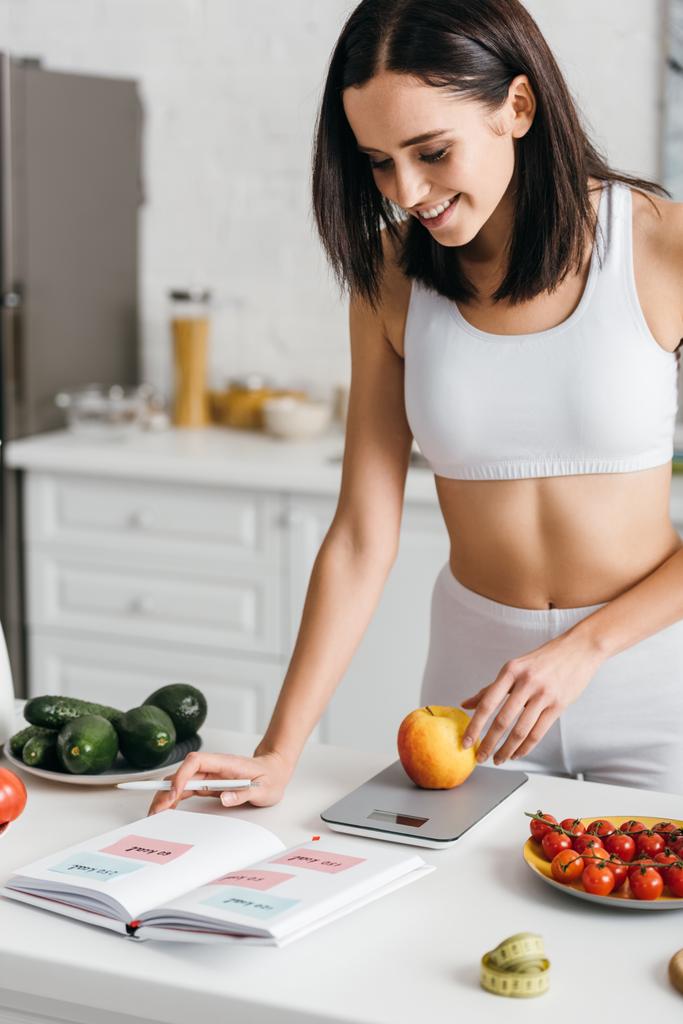 Selective focus of beautiful smiling sportswoman writing calories while weighing apple on kitchen table, calorie counting diet - 写真・画像