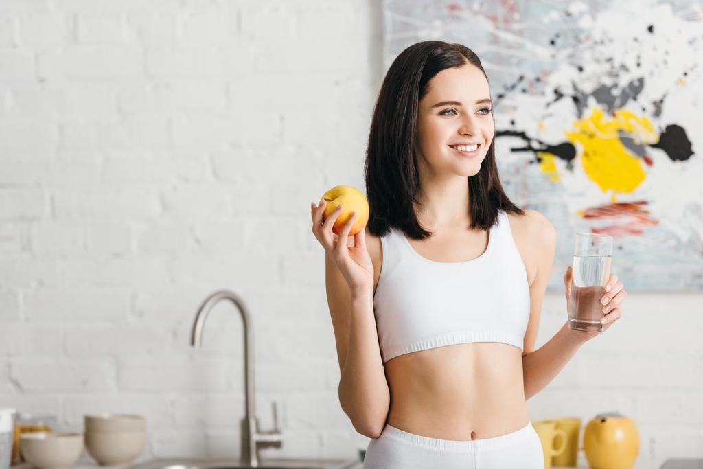 Sportive souriante tenant un verre d'eau et de pomme dans la cuisine
  - Photo, image