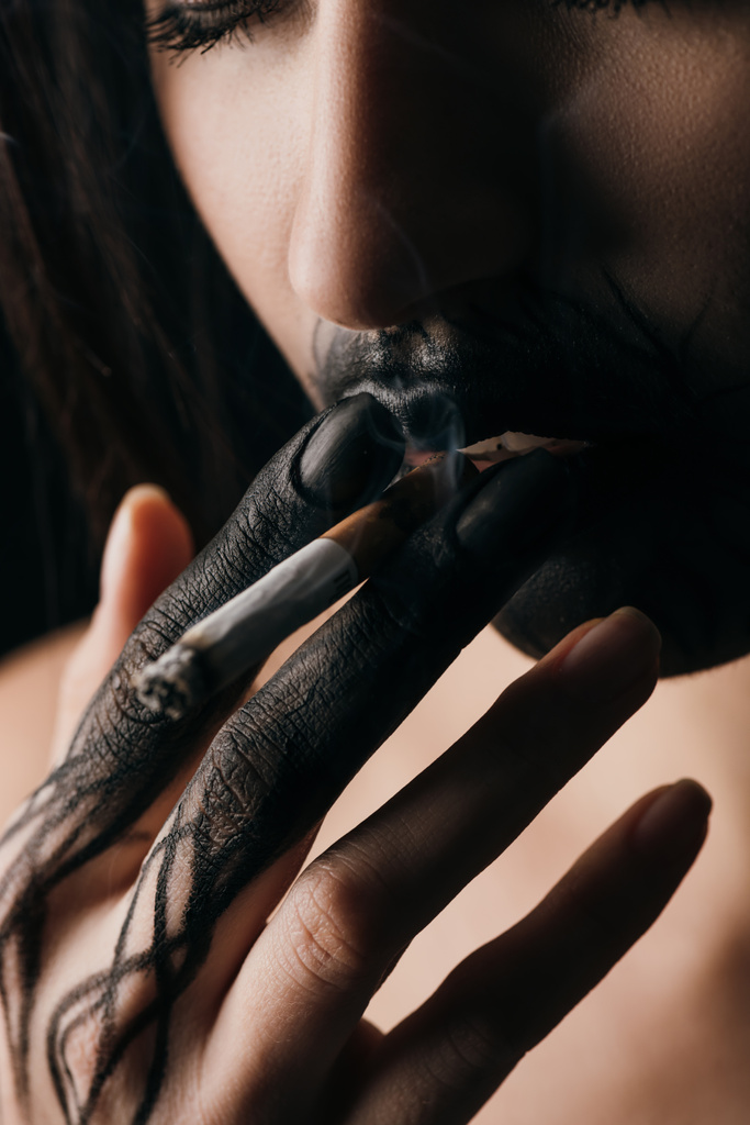 Selective focus of girl with black painted hand and lips smoking cigarette isolated on black  - Fotoğraf, Görsel