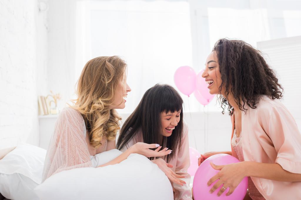 χαρούμενες πολυπολιτισμικές φίλες γελάνε στο κρεβάτι με ροζ μπαλόνια  - Φωτογραφία, εικόνα