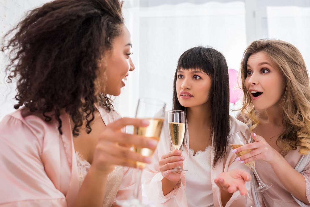 удивленные многонациональные девушки с бокалами шампанского, разговаривающие на пижамной вечеринке
 - Фото, изображение