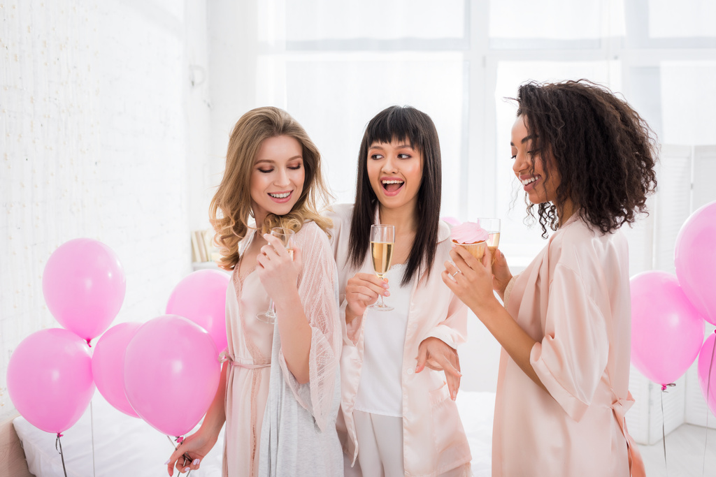счастливые мультиэтнические девушки веселятся с шампанским и кексом во время девичника с розовыми шариками
 - Фото, изображение