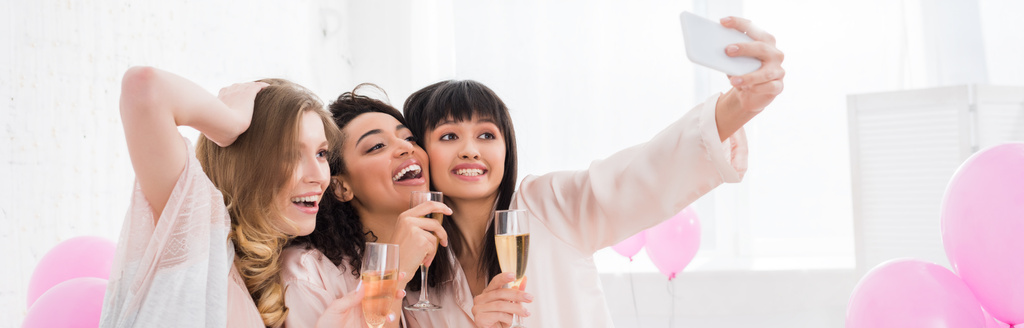 panoramic shot of emotional multicultural girls with glasses of champagne taking selfie on smartphone during pajama party - Photo, Image