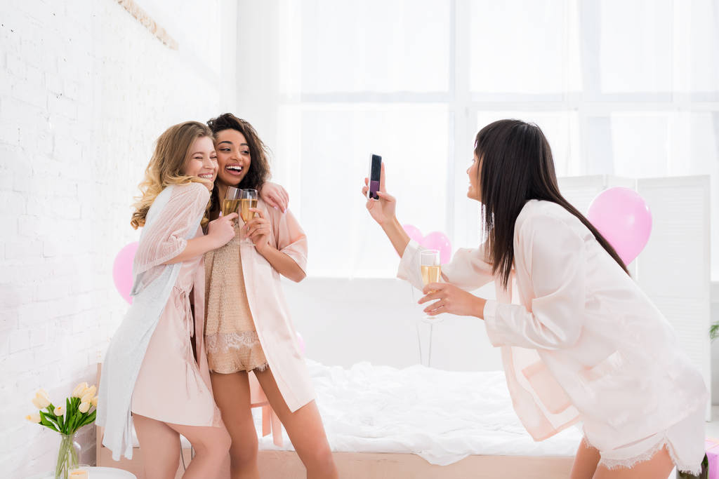 beautiful girl taking photo of multicultural happy girlfriends with glasses of champagne on smartphone during pajama party - Photo, Image
