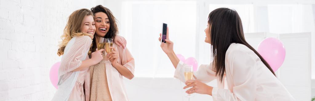 Panoramaaufnahme eines fröhlichen Mädchens, das während einer Pyjama-Party multikulturelle Freundinnen mit Champagnergläsern auf dem Smartphone fotografiert - Foto, Bild