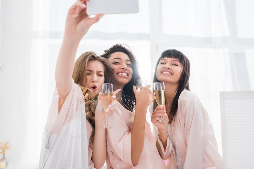 novias multiétnicas emocionales con copas de champán tomando selfie en el teléfono inteligente durante la fiesta de pijama
 - Foto, imagen