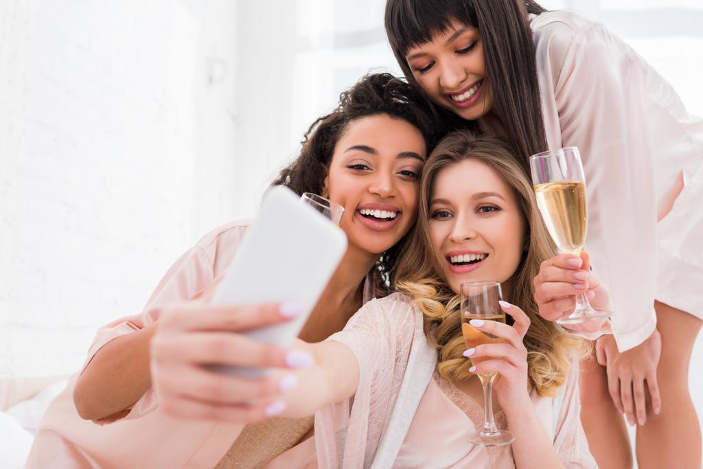 Gülümseyen, şampanya kadehli kızlar pijama partisi sırasında akıllı telefondan selfie çekiyorlar. - Fotoğraf, Görsel