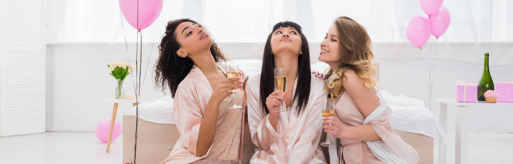 plano panorámico de amigas multiculturales positivas divirtiéndose y sosteniendo copas de champán en fiesta de pijama con globos rosados
 - Foto, imagen
