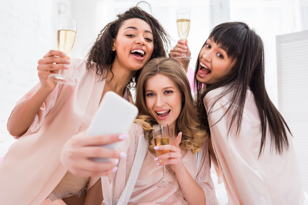 meninas multiculturais alegres com copos de champanhe tomando selfie no smartphone durante a festa de pijama
 - Foto, Imagem