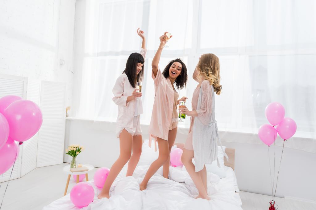 веселые мультиэтнические девушки танцуют с бокалами шампанского на девичнике с воздушными шарами
 - Фото, изображение