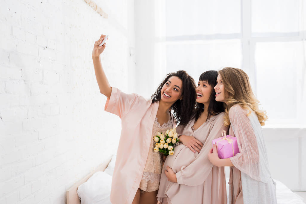 sonrientes novias multiculturales y feliz mujer embarazada sosteniendo flores y presente mientras toma selfie en baby shower
  - Foto, imagen