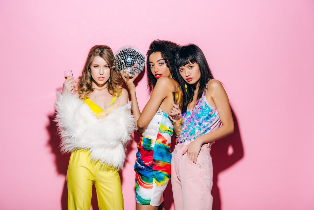 namoradas multiétnicas emocionais com champanhe e bola de discoteca posando em rosa
 - Foto, Imagem