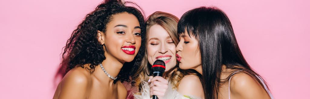Panoramaaufnahme fröhlicher multikultureller Mädchen, die Karaoke mit Mikrofon singen, isoliert auf rosa - Foto, Bild