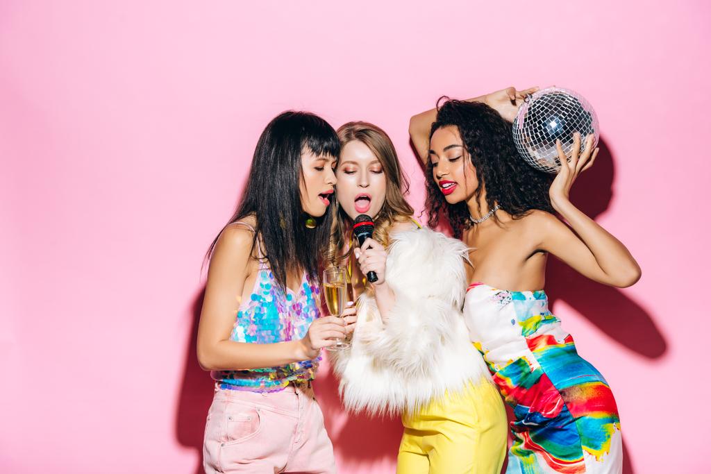 emotionele multiculturele vriendinnen die champagne en discobal vasthouden terwijl ze zingen met microfoon op roze - Foto, afbeelding
