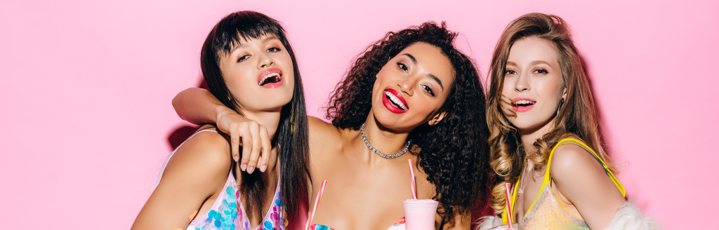 панорамный снимок счастливых модных мультиэтнических девушек, держащих стаканы с молочными коктейлями на розовом
 - Фото, изображение
