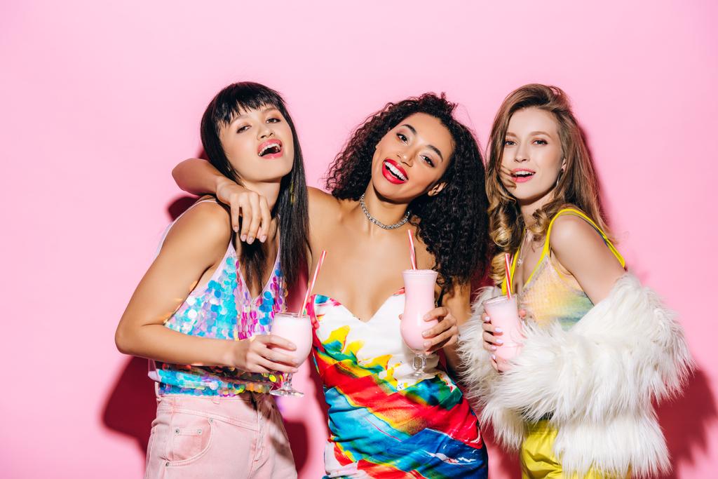 glimlachende stijlvolle multi-etnische meisjes houden bril met milkshakes op roze - Foto, afbeelding