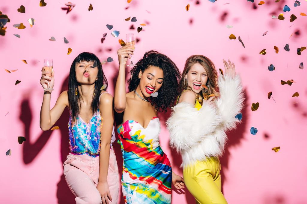 aufgeregte multikulturelle Mädchen amüsieren sich mit Sektgläsern auf rosa mit Konfetti - Foto, Bild