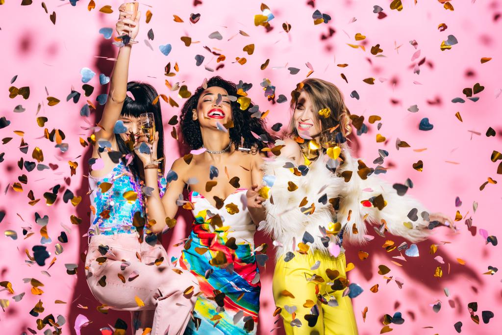 meninas multiculturais felizes se divertindo com copos de champanhe em rosa com confete
 - Foto, Imagem