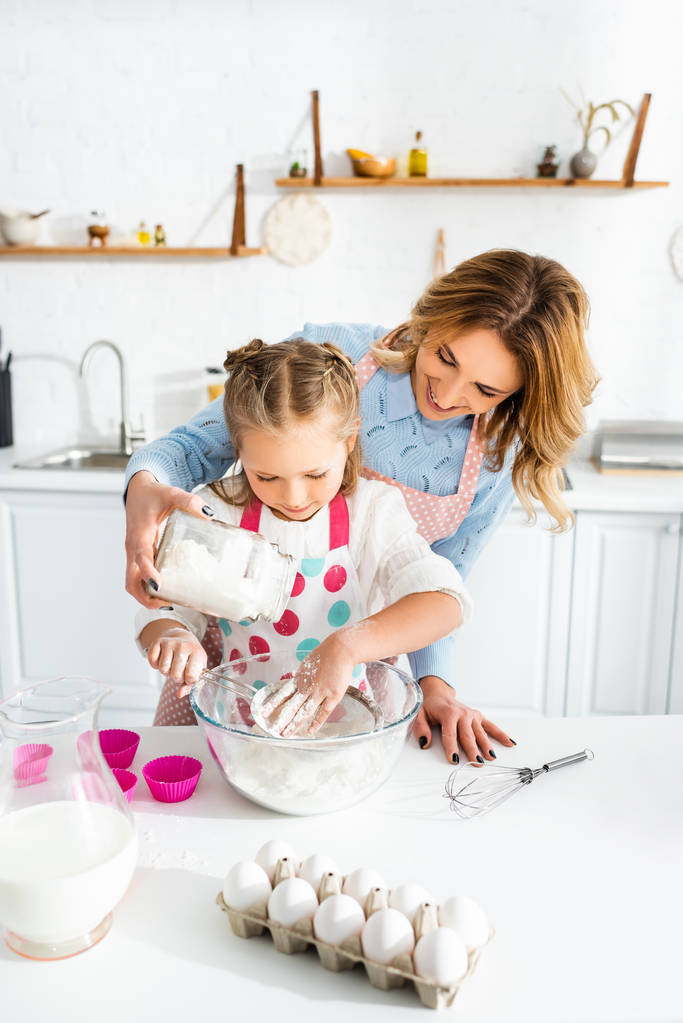 Belle mère et jolie fille tamiser la farine dans un bol sur la table près de moules à pâte, oeufs, cruche de lait et fouet de ballon
 - Photo, image