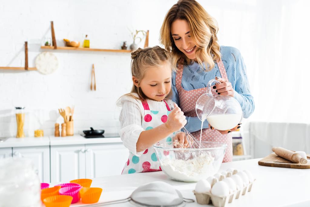 Selektywne skupienie córki ugniatanie ciasta, podczas gdy matka wylewa mleko z dzbanka w misce w domu - Zdjęcie, obraz