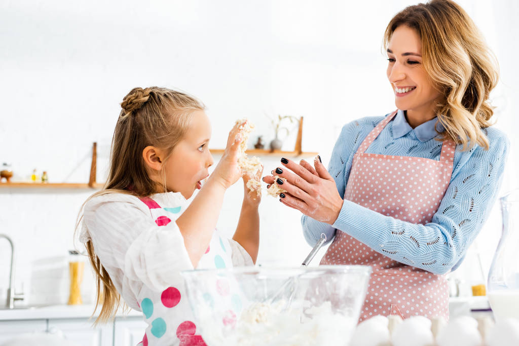 Selective focus of smiling mom and daughter with open mouth holding up hands having fun during kneading dough - Photo, Image