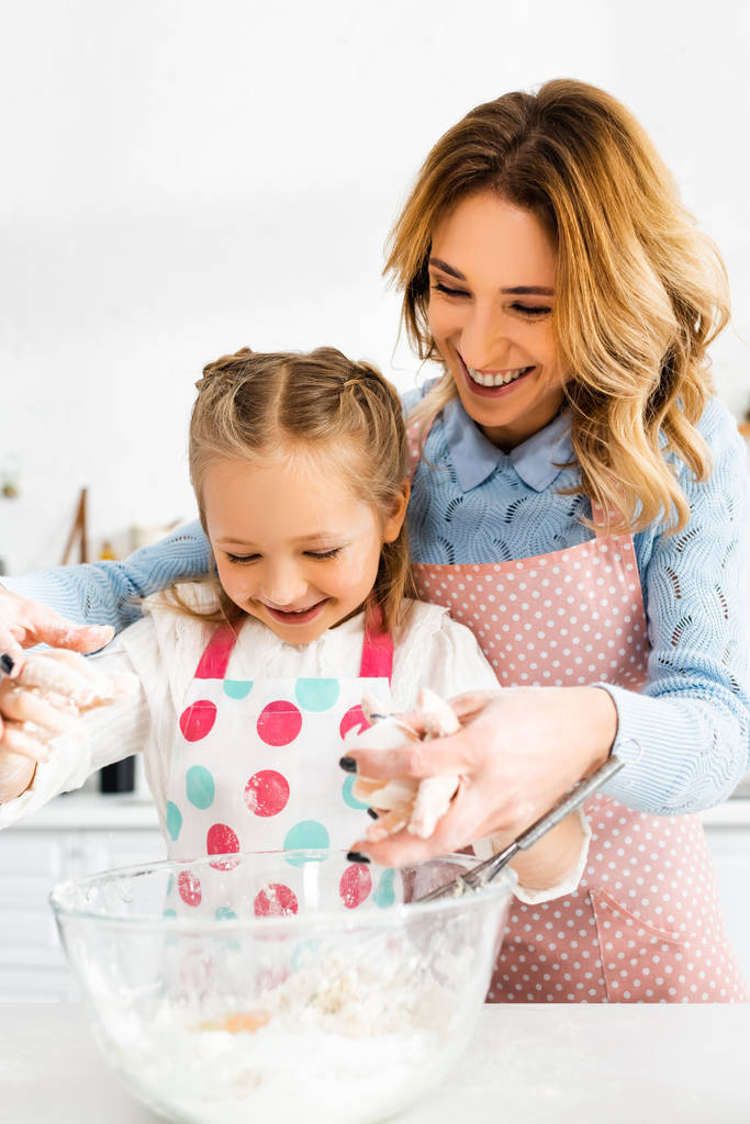 Селективный фокус привлекательной мамы и милой дочери улыбаясь и смешивая ингредиенты для вкусных кексов вместе
 - Фото, изображение
