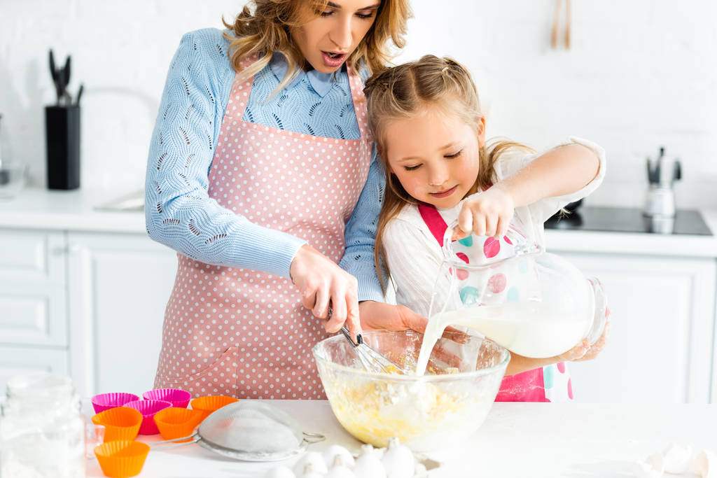 vista ritagliata della mamma che cucina con frusta palloncino e figlia versando latte
  - Foto, immagini