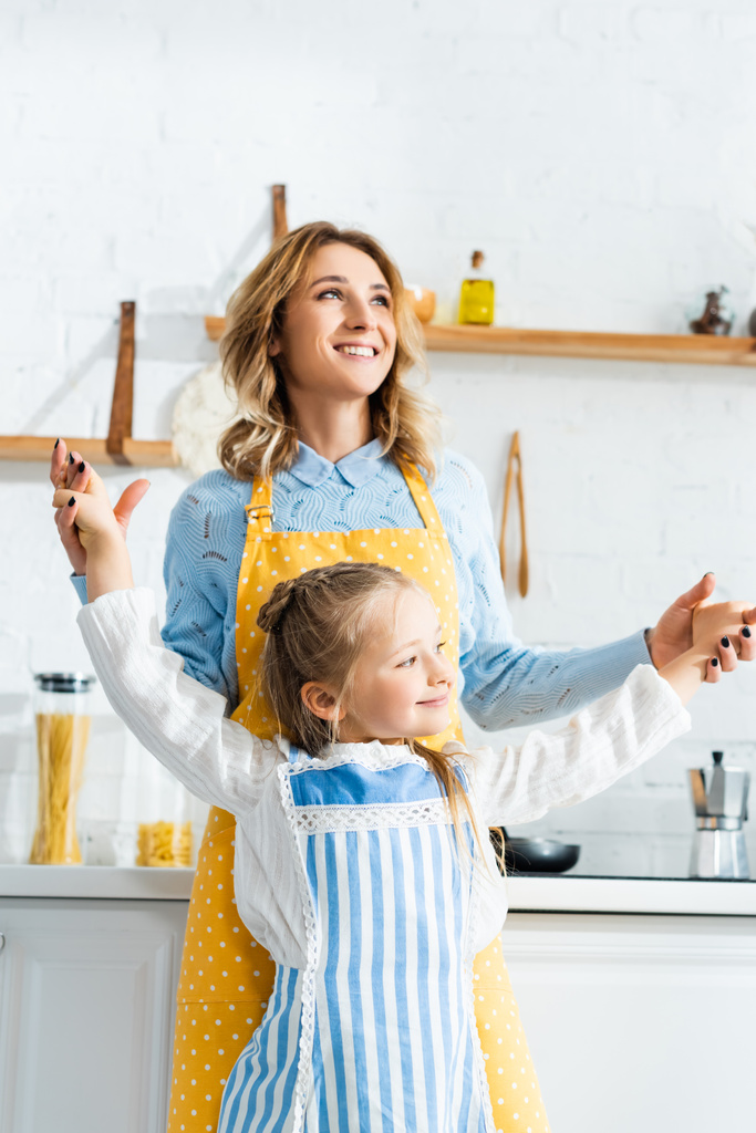 sonriente madre cogida de la mano con su hija y mirando a la cámara en la cocina
  - Foto, Imagen