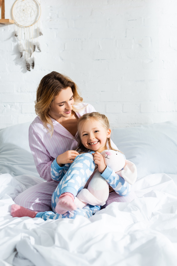 hymyilevä äiti ja söpö tytär pyjamassa istuu sängyssä
  - Valokuva, kuva