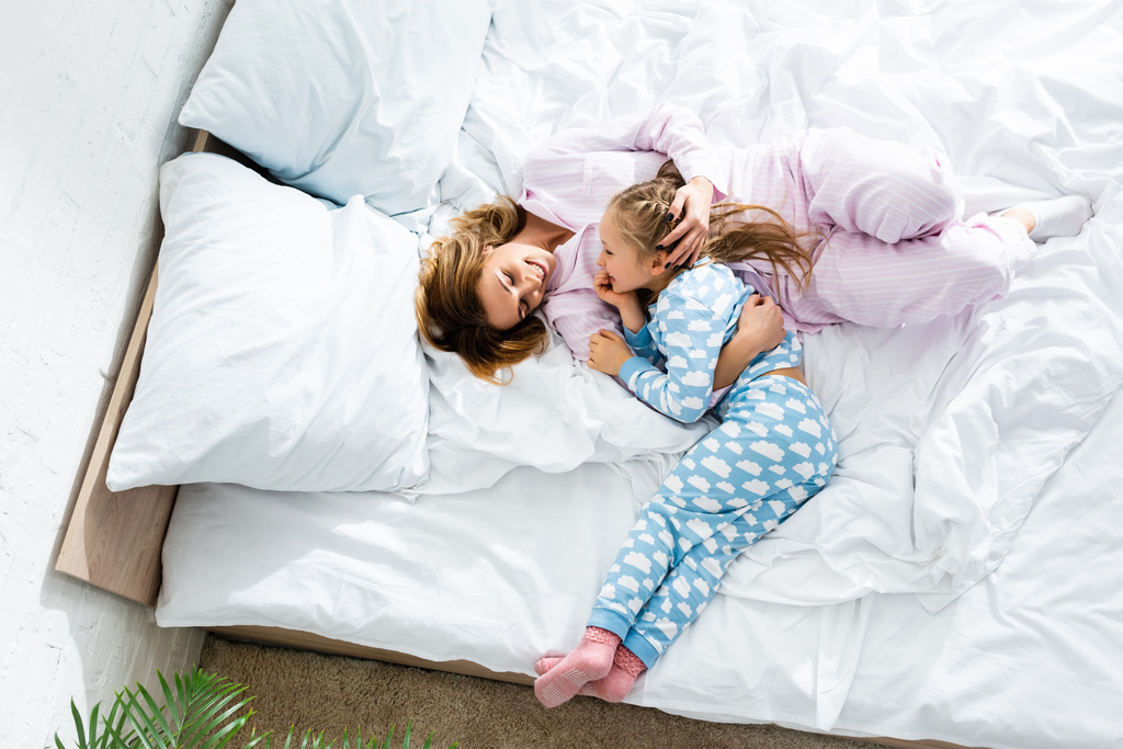 Вид сверху улыбающейся матери, обнимающей дочь и лежащей в постели
  - Фото, изображение