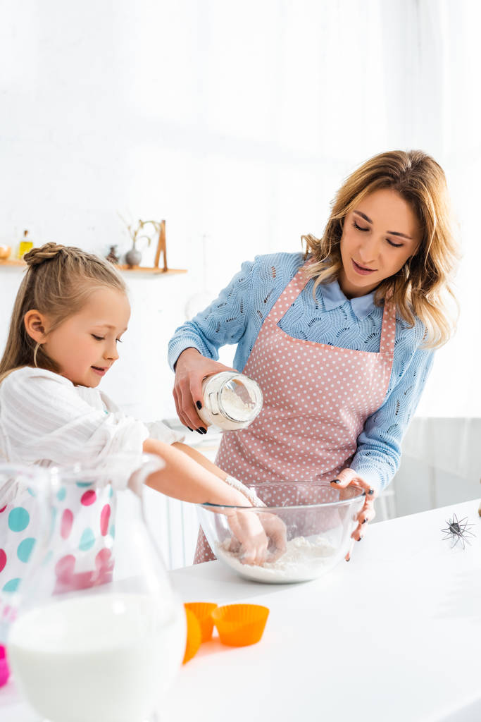 madre añadiendo harina al tazón y sonriente hija cocinando en la cocina
  - Foto, imagen