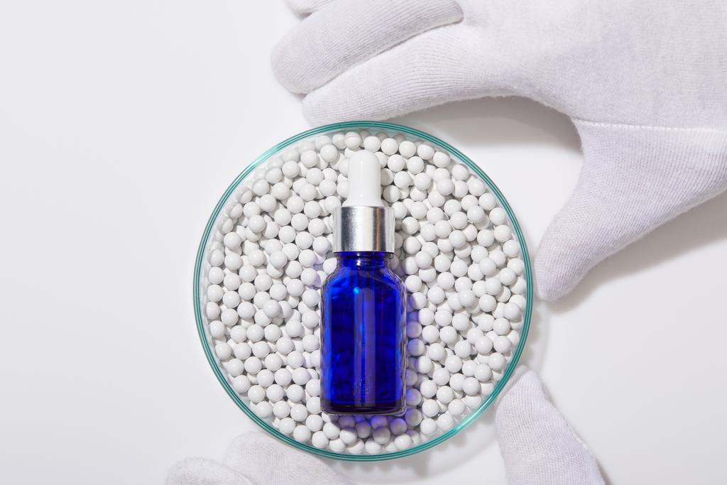 Cropped view of man holding laboratory glassware with blue bottle of oil on decorative beads on white background - Photo, Image
