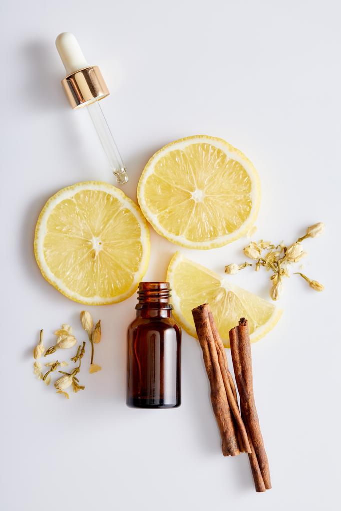 Vue du dessus de la bouteille d'huile cosmétique avec compte-gouttes, tranches de citron, bâtonnets de cannelle et bourgeons de vanille sur fond blanc
 - Photo, image